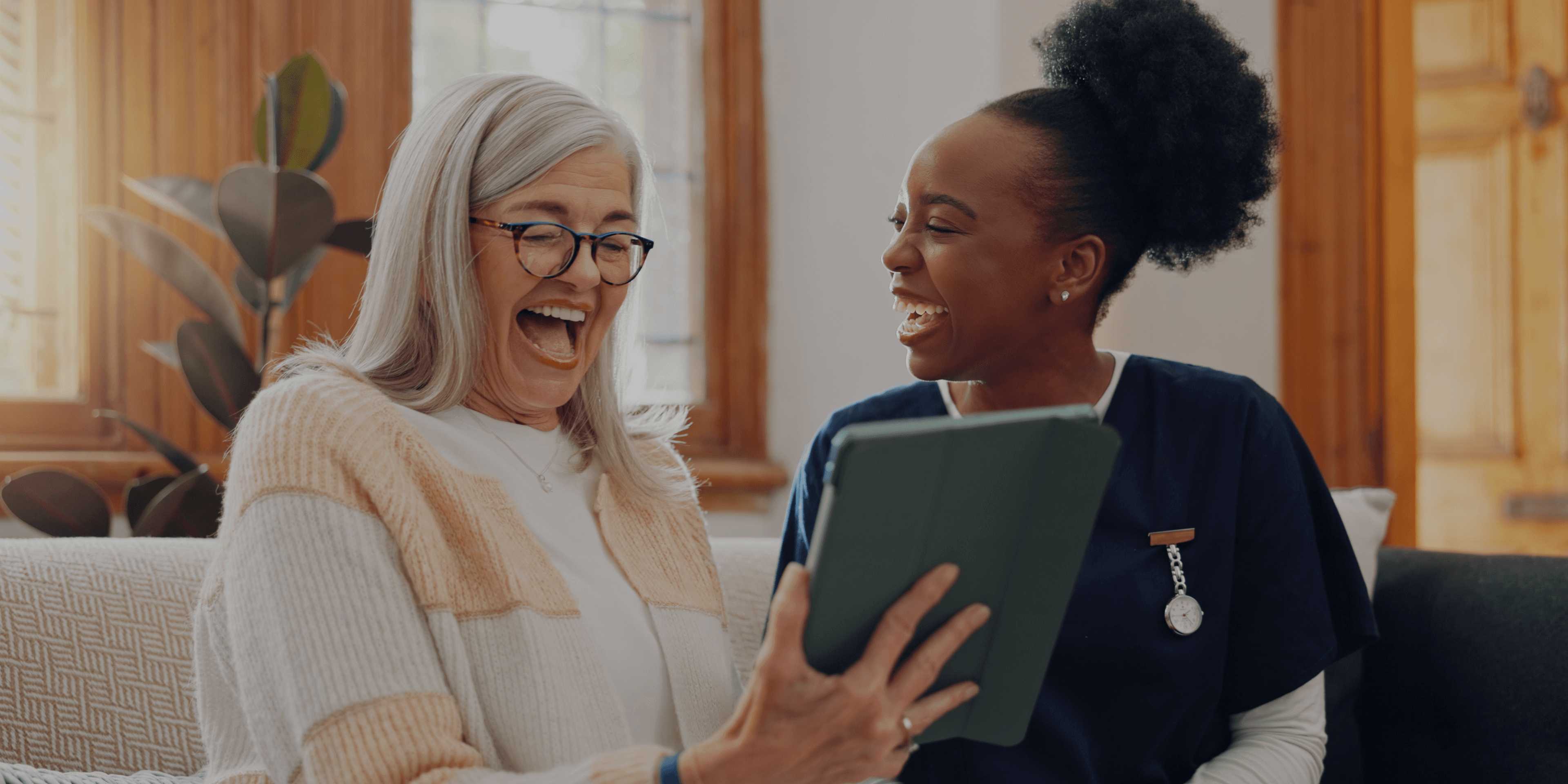Vrouw op Ipad aan het lachen met thuiszorgmedewerker naast zich op de bank